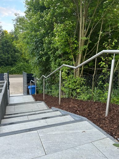 Treppen mit Handlauf, umgeben von Bäumen und mulchbedecktem Boden.